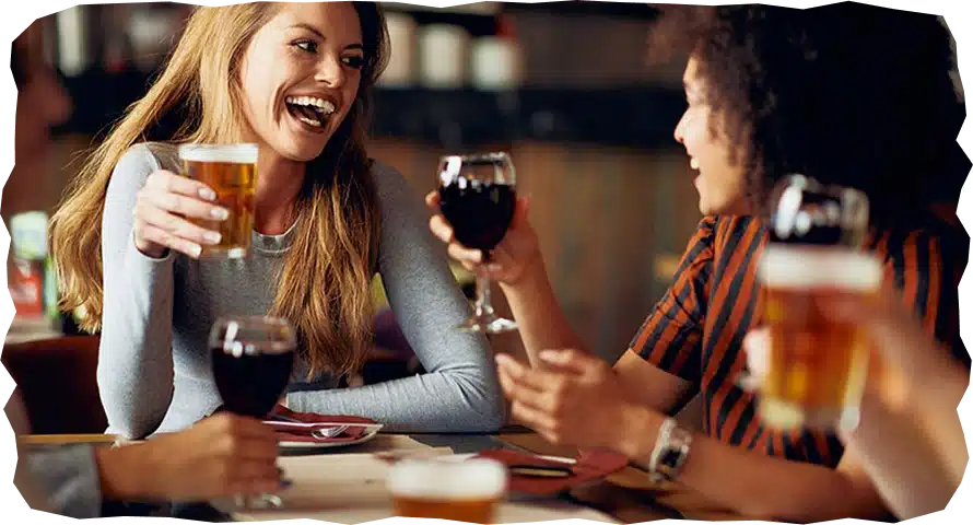 Two women sit at a table in a lively bar, smiling and laughing as they toast with beer and wine. The friendly vibe is perfect to plan your visit to Strandhill Surf School after a fun day on the waves.