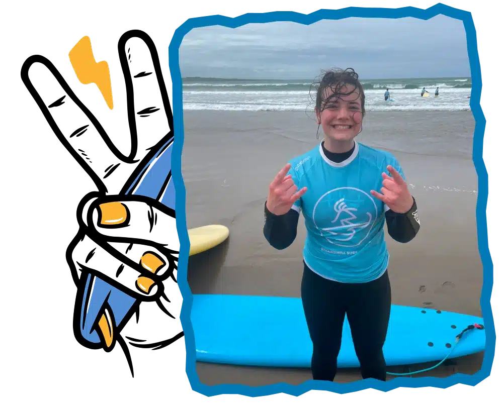A smiling surfer in a blue rash vest and black wetsuit stands on Strandhill Beach after Surf Lessons Strandhill Sligo, making a "rock on" gesture. A blue surfboard lies behind them as the sea and fellow surfers fill the background.