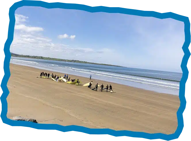 A group of people with surfboards stands on a wide sandy beach near the water’s edge under a blue sky, enjoying gentle waves and the distant coastline—an inviting scene framed by a jagged blue border for Strandhill Surf Lessons.