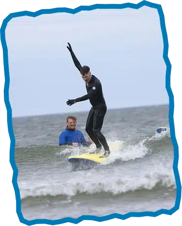 A person in a black wetsuit balances on a yellow surfboard, riding a small wave, as another in blue watches from the water—a classic scene from Private Surf Lessons Strandhill. The image features a jagged blue border.