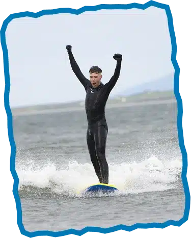 A person in a black wetsuit stands upright on a surfboard, raising both arms in triumph whilst riding a small wave—perfect for Intermediate Surf Lessons Strandhill. The background shows a cloudy sky and distant land. The image has a blue border.