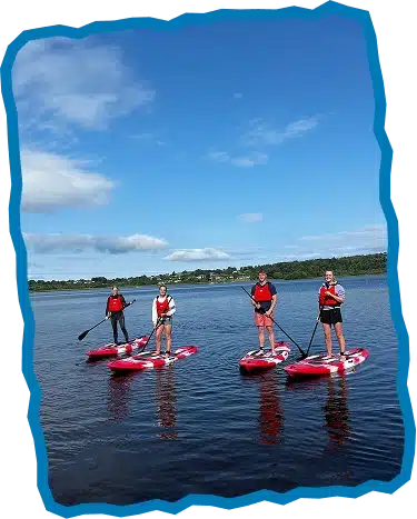 Four people wearing red life jackets stand on paddleboards on a calm lake under a blue sky with scattered clouds, enjoying an experience similar to Surf Lessons Strandhill Sligo. Green trees and houses line the distant shoreline.