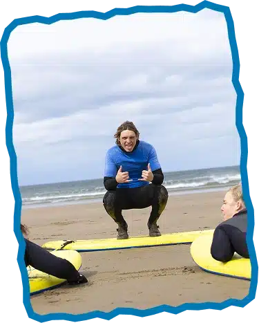 A person in a blue shirt and wetsuit squats on the sand by a yellow surfboard, giving a thumbs up during Children’s Surf Lessons Strandhill, whilst two others with surfboards listen, with the sea and cloudy sky in the background.