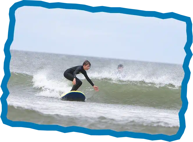 A child in a black wetsuit surfs a small wave on a blue board during Strandhill Surf Lessons, balancing skilfully as another person watches from the water. The scene is framed by a wavy blue border.