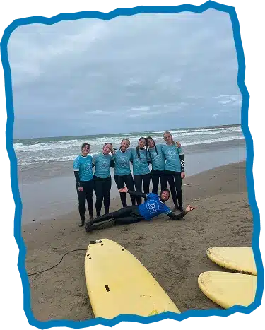 Six people in wetsuits and blue shirts smile together on a sandy beach with waves in the background at Surf Lessons Strandhill Sligo. One person lies playfully in front whilst two yellow surfboards rest nearby.