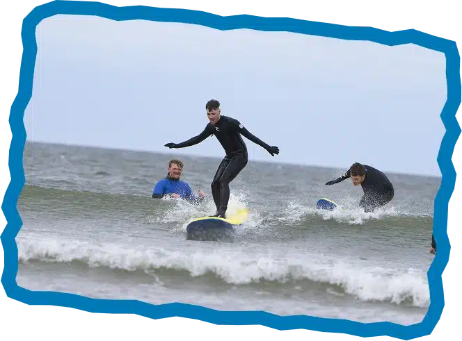Three people in wetsuits surf small waves in the sea under a cloudy sky during Hen and Stag Surf Lessons Strandhill. One stands on a yellow surfboard, while the others balance nearby. A wavy blue border frames the photo.