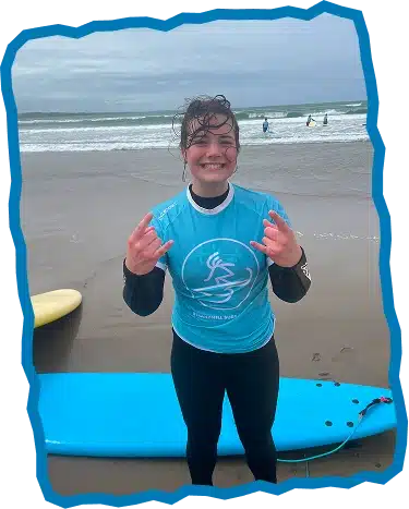 A smiling person in a blue surf shirt and black wetsuit stands on a sandy beach at Surf Lessons Strandhill Sligo, making the "rock on" hand gesture. A blue surfboard lies nearby, with waves and cloudy skies in the background.