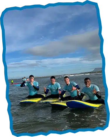 Four men in light blue rash vests sit on surfboards in shallow sea water, smiling and making "shaka" hand signs. Enjoying Surf Lessons Strandhill Sligo, they relax as waves roll in under a partly cloudy sky with land in the distance.