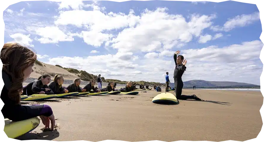 A group of people in wetsuits sit on surfboards on a sandy beach, attentively watching an instructor from Strandhill Surf School demonstrating a surfing move under a partly cloudy sky, with sand dunes and hills in the background.