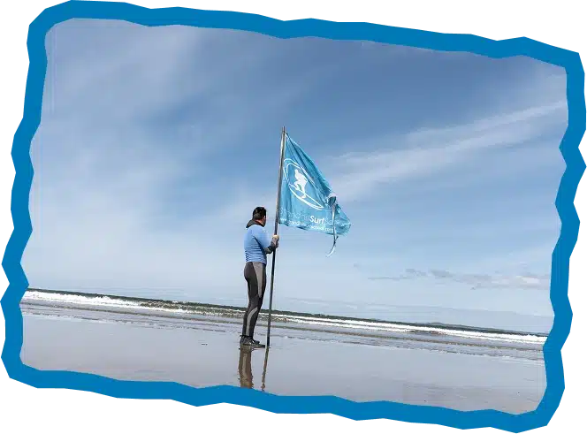 A person in a wetsuit and blue shirt stands on a sandy beach, holding a blue flag with a white surf logo—perfect for promoting Strandhill Surf Lessons. The ocean waves and a partly cloudy sky are framed by a wavy blue border.