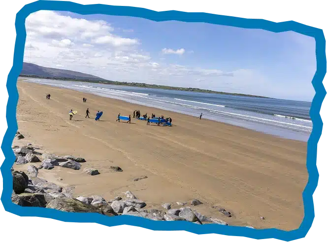 A group of people on a wide sandy beach near the water, some carrying blue surfboards at Strandhill Summer Surf Camp. The sky is partly cloudy and waves gently roll in; rocks are in the foreground with grassy hills beyond.