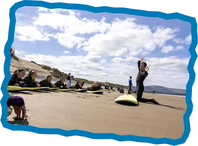 A surf instructor in a wetsuit demonstrates techniques on the sand to a group of students sitting on surfboards at a sunny beach, with dunes and the sea in the background during Surf Lessons Strandhill Sligo.