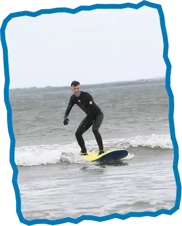 A person wearing a black wetsuit enjoys surfing a small wave on a yellow and blue surfboard, under a grey sky with the distant shoreline behind—capturing the spirit of Private Surf Lessons Strandhill.