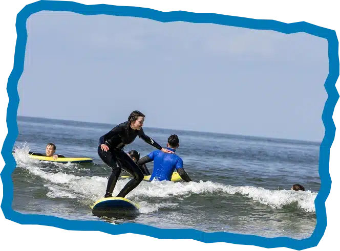 A group of people in wetsuits enjoy small waves together on the sea, with one person surfing in the foreground and others paddling nearby under a partly cloudy sky—perfect for Surf Camps Strandhill. A blue border frames the image.