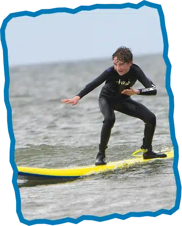A young person in a wetsuit balances on a yellow surfboard, riding a small wave with focused determination during Children’s Surf Lessons Strandhill. The ocean scene is framed by a jagged blue border.