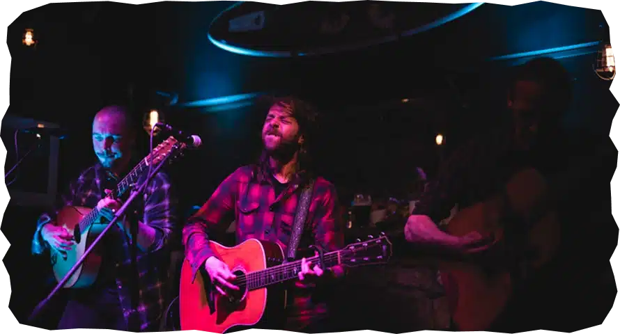 Three musicians wearing checked shirts play acoustic guitars on a dimly lit stage, illuminated by purple and blue lighting, creating a moody, intimate atmosphere—perfect for those looking to explore Strandhill Sligo’s vibrant live music scene.