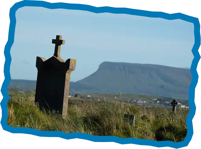 A weathered gravestone with a cross stands in grassy terrain, overlooking distant houses and a mountain under a clear blue sky. Explore Strandhill Sligo, where even the smallest cross-shaped markers tell tales of history.