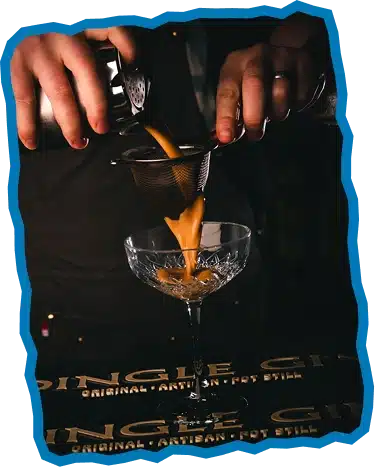 A person pours a creamy cocktail from a shaker through a strainer into a crystal glass, their hands and the drink in focus against a dark background—capturing the refined spirit you’ll find when you explore Strandhill Sligo.