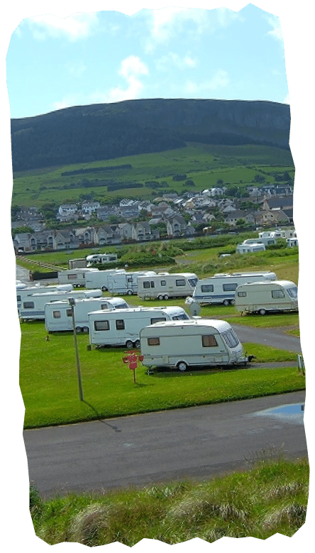 Rows of white caravans are parked on green grass near a small town, with rolling hills and a cloudy sky in the background—an ideal spot to plan your visit to Strandhill Surf School.